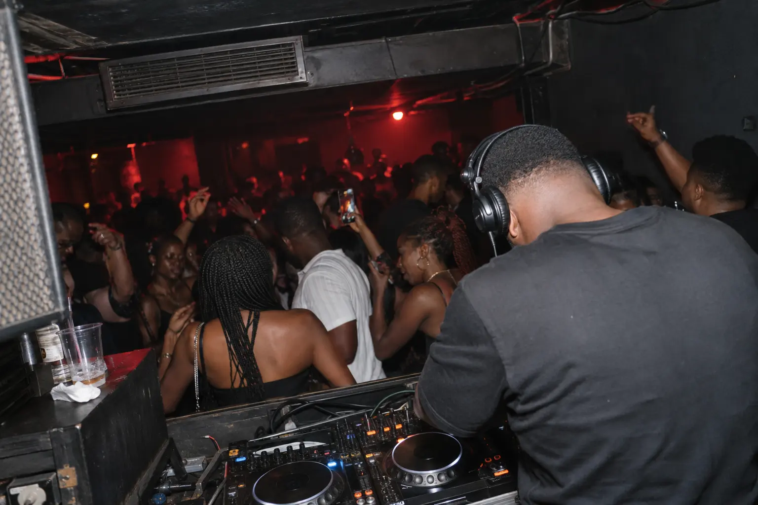 A DJ seen from behind the decks, facing a full dancefloor lit red